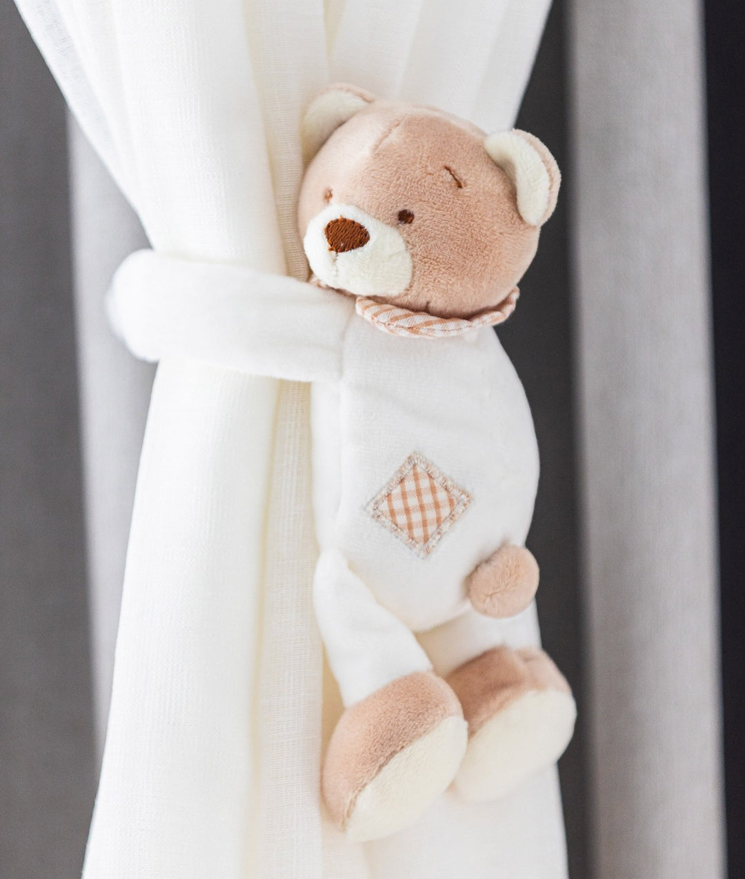 Beige and white plush teddy bear curtain tieback with embroidered face and gingham tummy holding sheer curtain