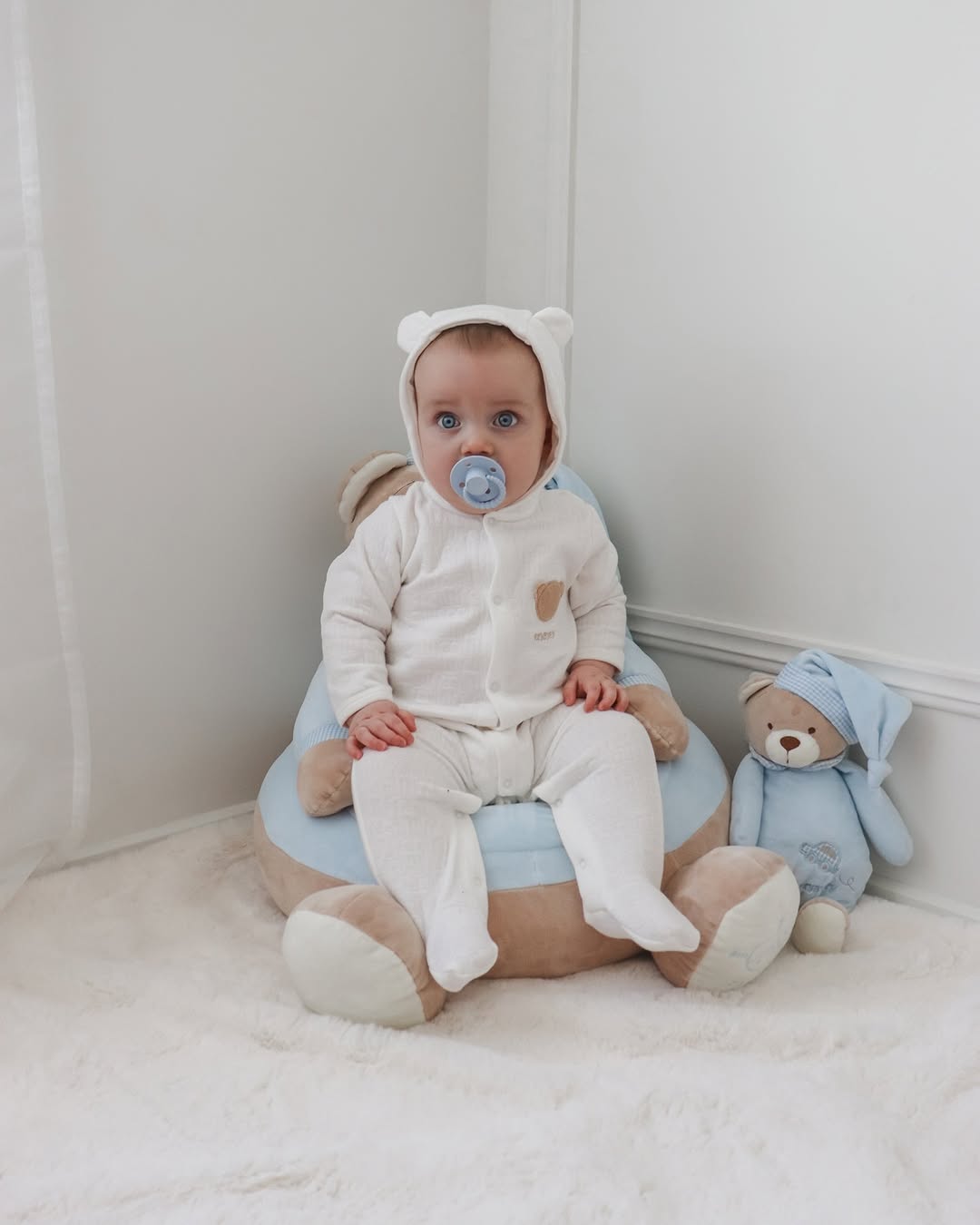 Baby wearing white hooded bear jumpsuit sitting on plush blue cushion with hood up, showcasing cozy nursery use case