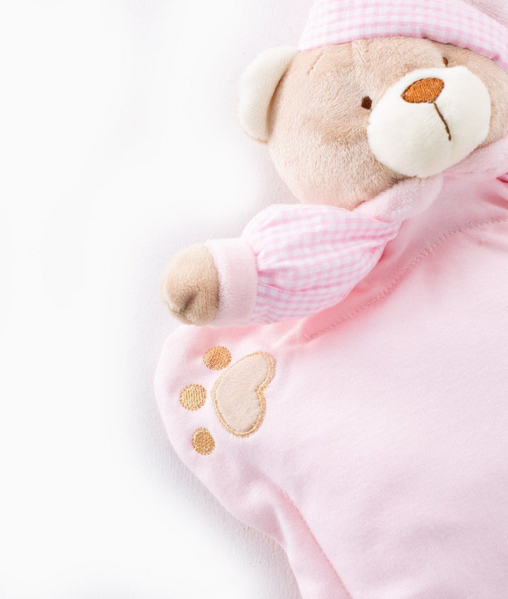 Plush light brown teddy bear wearing pink gingham hat resting on soft solid pink cotton fabric